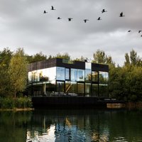 Foto: Pasakaina stikla māja uz ezera, kurā atspoguļojas apkārtnes ainava